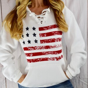 May include: White hooded sweatshirt with a distressed American flag design. The flag features red stripes and black stars. The sweatshirt has a lace-up neckline and a front pocket.