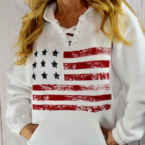 May include: White hooded sweatshirt featuring a distressed American flag design. The flag graphic includes red stripes and black stars. The sweatshirt has a lace-up neckline and a front pocket.