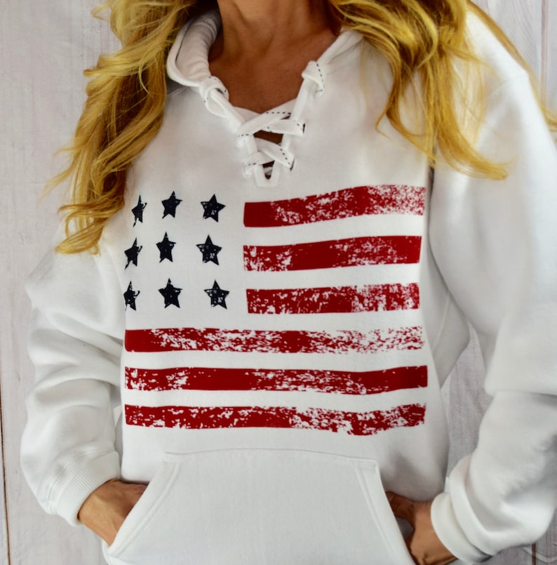 May include: White hooded sweatshirt featuring a distressed American flag design. The flag graphic includes red stripes and black stars. The sweatshirt has a lace-up neckline and a front pocket.