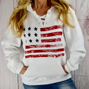May include: White hooded sweatshirt featuring a distressed American flag design. The flag graphic includes red stripes and black stars. The sweatshirt has a lace-up neckline and a front pocket, perfect for casual wear.