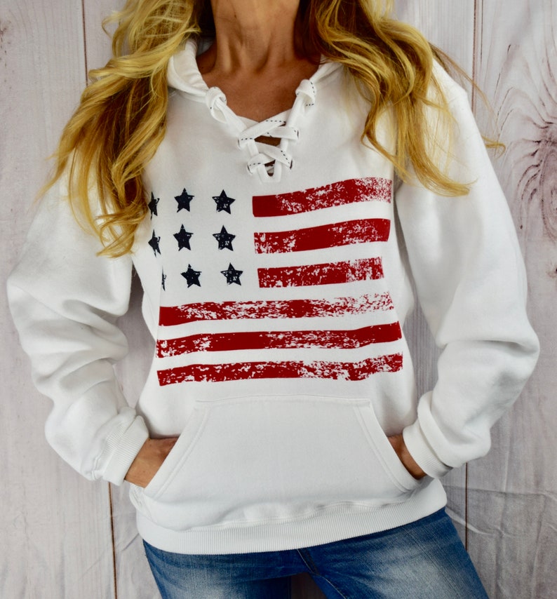 May include: White hooded sweatshirt featuring a distressed American flag design. The flag graphic includes red stripes and black stars. The sweatshirt has a lace-up neckline and a front pocket, perfect for casual wear.