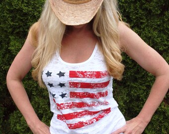 May include: A woman wearing a white tank top with a distressed American flag design and blue jeans. She is wearing a straw cowboy hat.