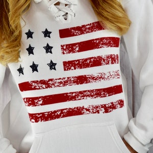 May include: White hooded sweatshirt featuring a distressed American flag design in red and navy blue. The sweatshirt has a lace-up neckline and a front pocket. The design includes red stripes and navy blue stars.