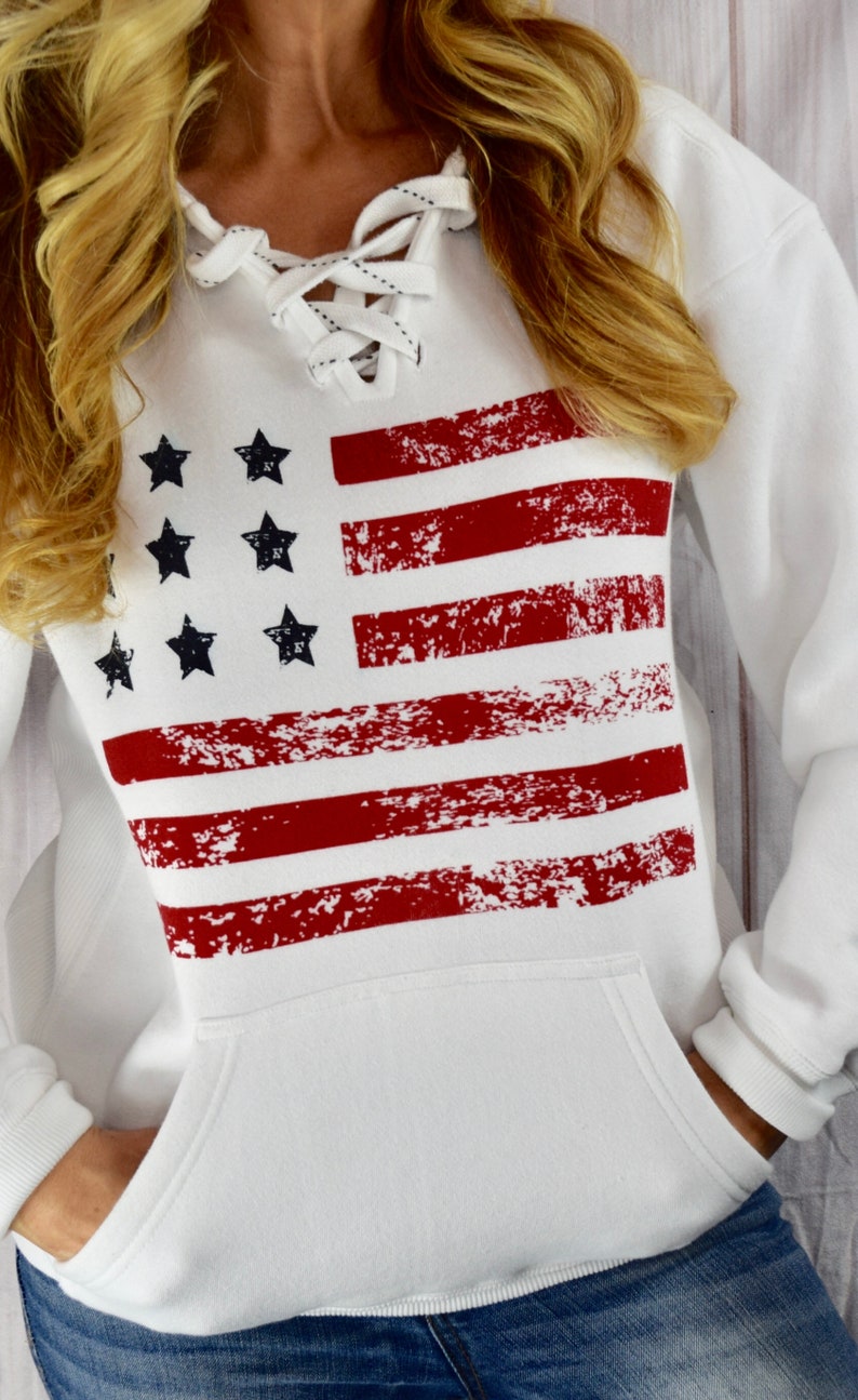 May include: White hooded sweatshirt featuring a distressed American flag design in red and navy blue. The sweatshirt has a lace-up neckline and a front pocket. The design includes red stripes and navy blue stars.