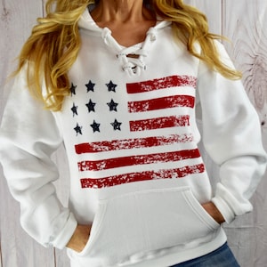 May include: White hooded sweatshirt with a distressed American flag design. The flag features red stripes and black stars. The sweatshirt has a lace-up neckline and a front pocket.