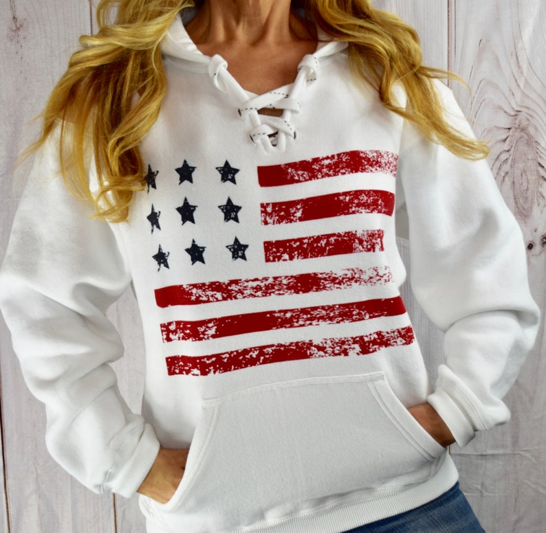 May include: White hooded sweatshirt with a distressed American flag design. The flag features red stripes and black stars. The sweatshirt has a lace-up neckline and a front pocket.