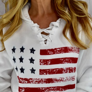 May include: White hooded sweatshirt featuring a distressed American flag design. The flag has red stripes and navy blue stars. The neckline has a lace-up detail. The sweatshirt is a casual, patriotic apparel item.