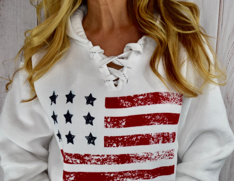 May include: White hooded sweatshirt featuring a distressed American flag design. The flag has red stripes and navy blue stars. The neckline has a lace-up detail. The sweatshirt is a casual, patriotic apparel item.