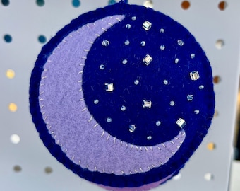 May include: A purple felt crescent moon with a blue felt background decorated with silver glitter stars. This handmade ornament is perfect for a nursery or child's room.