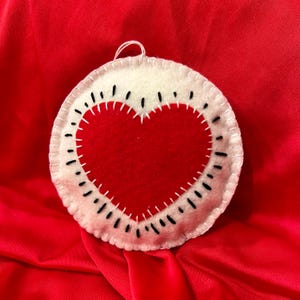 May include: A white felt circle with a red felt heart in the center. The heart is stitched with black thread and the circle is stitched with white thread.