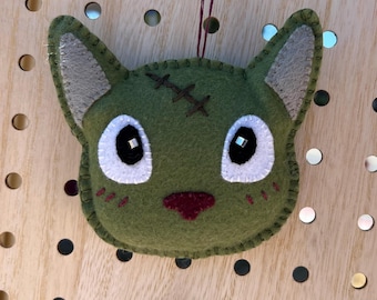 May include: A handmade green felt cat ornament with stitched details. The cat has white eyes with black pupils, a red nose, and stitched details. The ears are grey, and the head features stitched lines. A string is attached for hanging.