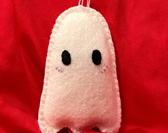 May include: A white felt ghost decoration with black button eyes and pink stitched cheeks. The ghost has a stitched outline and a white string for hanging. The background is a red fabric.