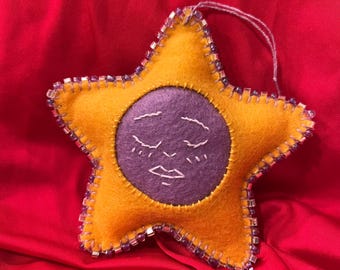 May include: A handmade star-shaped ornament with a yellow felt exterior and a purple felt circle in the centre. The purple circle features a stitched face with closed eyes and a small mouth. The edges are embellished with beads.