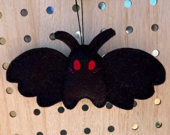 May include: A black felt moth ornament with red eyes and a cute expression. The moth is hanging from a string.