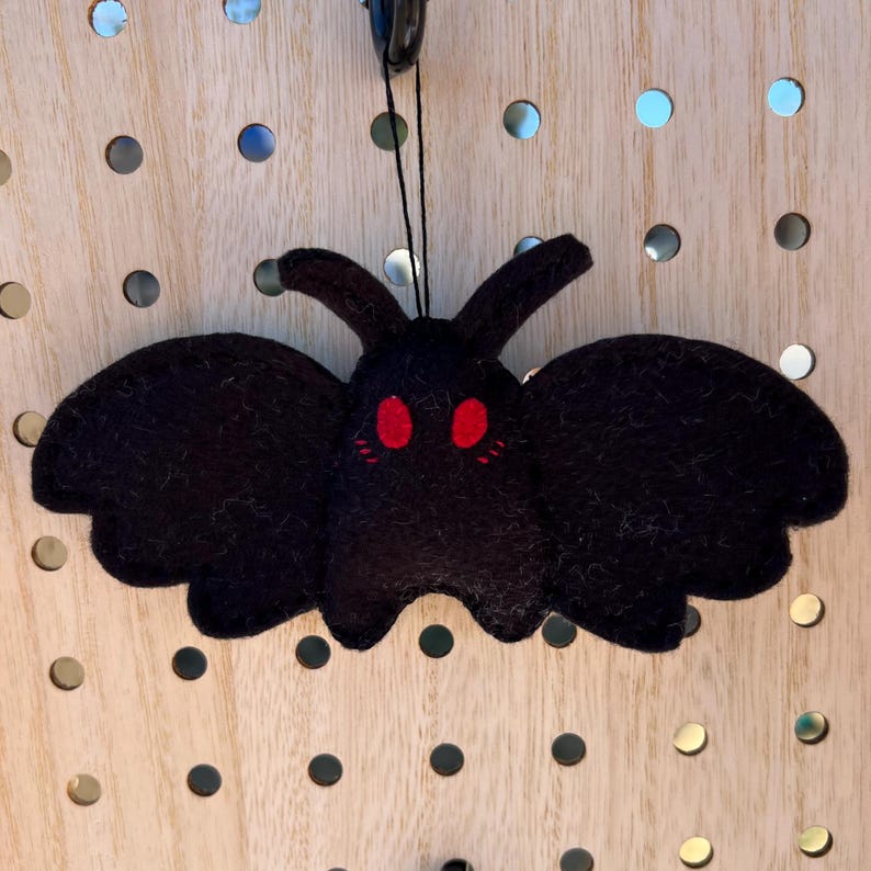 May include: A black felt moth ornament with red eyes and a cute expression. The moth is hanging from a string.