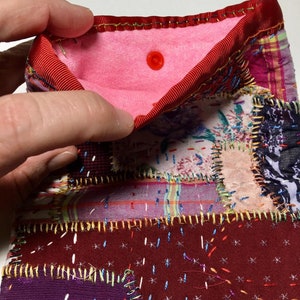 Puede incluir: Una bolsa de tela de retazos con un ribete de cinta roja y un interior rosa. La bolsa está hecha de varios retazos de tela cosidos con hilo dorado y colorido. Un botón rojo asegura la solapa.