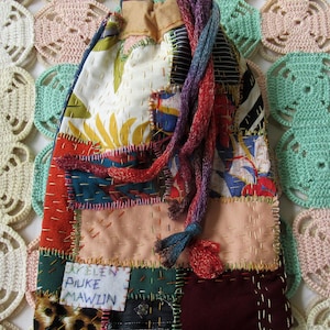 Puede incluir: Un bolso con cordón de patchwork con una variedad de telas en tonos rojos, verdes, azules y crema. El bolso presenta un estampado floral, de leopardo y patrones de rayas. El cordón es un cordón trenzado de hilos morados, azules y rojos. El bolso tiene una etiqueta que dice "AYELEN PIUKE MAWLIN".