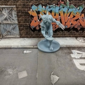 Peut inclure: Une figurine miniature en plastique gris d'un super-héros avec des griffes, portant un costume avec une ceinture et une cape. La figurine est debout sur une base grise.