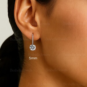 May include: A single silver earring with a round, clear gemstone, approximately 5mm in diameter, is shown in this image. The earring is suspended from a thin, curved silver hook. The earring is worn on a person's ear.