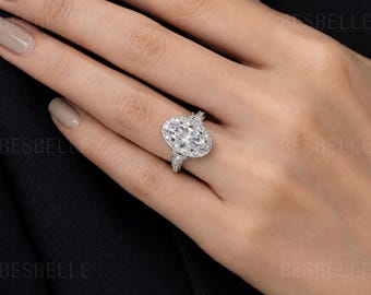May include: A silver-toned ring featuring a large oval-cut center stone surrounded by a halo of smaller round stones, set on a band accented with additional small stones. The word BESBELLE is visible in the background of the image.