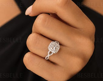 May include: A silver-toned ring featuring a large, cushion-cut center stone surrounded by a halo of smaller round stones. The band has a split, twisted design encrusted with additional small round stones. The word BESBELLE is visible.