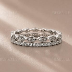 May include: A silver-toned ring set featuring two stacked bands. The top band has a series of marquise-shaped settings with small clear stones, while the bottom band is a thin eternity style with pavé-set stones. The inner band is marked 925.
