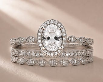 May include: A silver-toned ring set consisting of three stacked bands. The central band features an oval-cut clear stone surrounded by a halo of small round stones, while the two outer bands display a repeating marquise-shaped milgrain design.