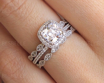Op de afbeelding: Een zilverkleurige verlovingsringset met een cushion-cut centrale steen omringd door een halo van kleine ronde steentjes. De ring en de bijbehorende stapelring zijn voorzien van gedetailleerde milgrain-afwerkingen en kleine steentjes.
