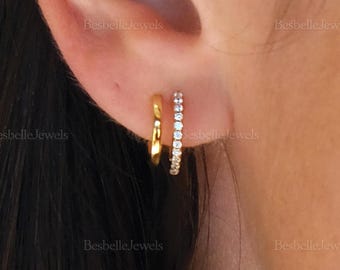 May include: A close-up of a person's ear with two gold hoop earrings. One earring is a plain gold hoop, and the other is a gold hoop with small, clear gemstones. The earrings are small and delicate.
