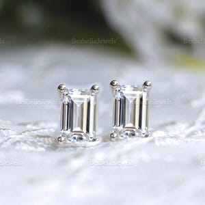 May include: A pair of square-cut, clear gemstone stud earrings. The earrings are set in a silver metal with a small, round accent at the top of each earring. The background is a soft, blurred white.