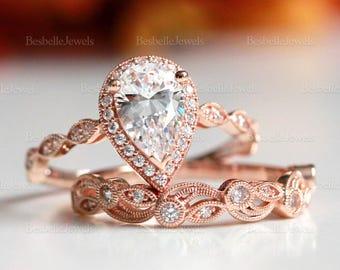 May include: Rose gold engagement ring set featuring a pear-shaped center stone surrounded by a halo of smaller stones. The matching band has a delicate, ornate design with additional small stones. The rings are set against a soft, blurred background.