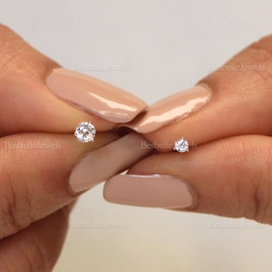 Op de afbeelding: Twee kleine, ronde, heldere diamant oorknopjes. De oorbellen worden tussen twee vingers gehouden met een manicure in neutrale tinten. De achtergrond is een zachte, neutrale kleur.