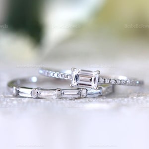 Op de afbeelding: Drie zilveren ringen met heldere edelstenen. Eén ring heeft een rechthoekig geslepen middensteen, terwijl een andere een rij kleinere stenen heeft. De derde ring heeft een reeks rechthoekige stenen. De ringen staan tegen een zachte, vage achtergrond.