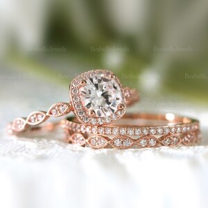 Puede incluir: Un juego de anillos de compromiso de oro rosa con un anillo de diamantes con halo cuadrado y dos bandas a juego. Los anillos están adornados con pequeños diamantes brillantes, creando un diseño delicado. El conjunto se muestra sobre una superficie blanca.