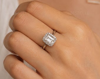 May include: A silver-toned ring featuring a central emerald-cut stone surrounded by a halo of smaller round stones. The band is thin and polished, showcasing a clear, faceted design that reflects light. The word BESBELLE is visible on the image.