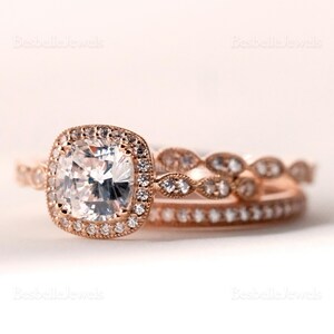 May include: A rose gold engagement ring set featuring a square-cut center stone surrounded by a halo of smaller stones. The set includes a matching band with delicate detailing and a second band with a row of small stones. The rings are set against a soft, neutral background.