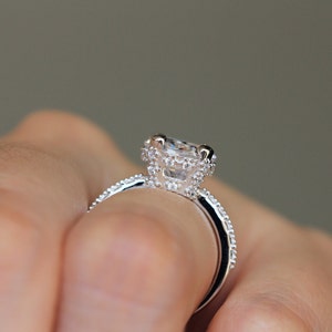 May include: Close-up of a silver ring with a large, clear, square-cut center stone. The ring features a halo of smaller stones around the center and along the band, creating a sparkling effect. The ring is held by a person.
