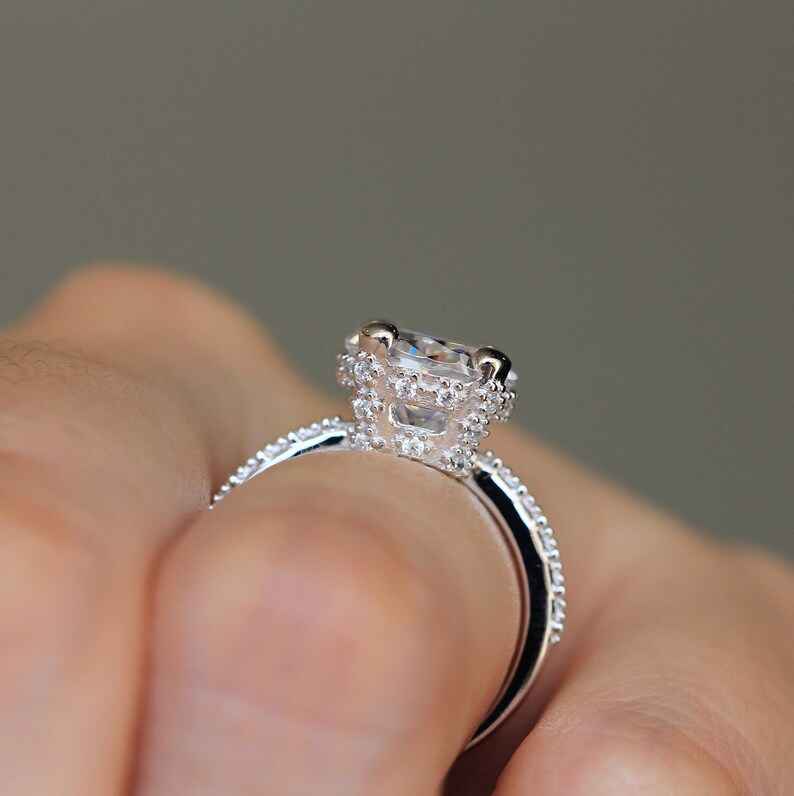 May include: Close-up of a silver ring with a large, clear, square-cut center stone. The ring features a halo of smaller stones around the center and along the band, creating a sparkling effect. The ring is held by a person.