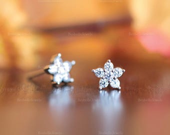 May include: A pair of small, flower-shaped stud earrings. Each earring features five clear, sparkling gemstones arranged in a floral pattern. The earrings are set on a post, and the background is a blurred mix of warm colors.