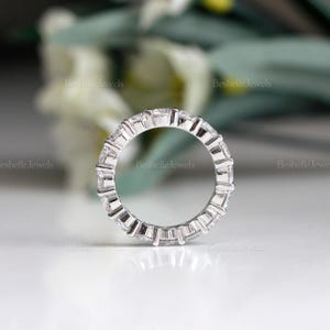 May include: A silver ring with a continuous row of square-cut, clear gemstones. The ring is set in a silver band and is displayed against a soft, blurred background. The ring is a piece of jewelry.