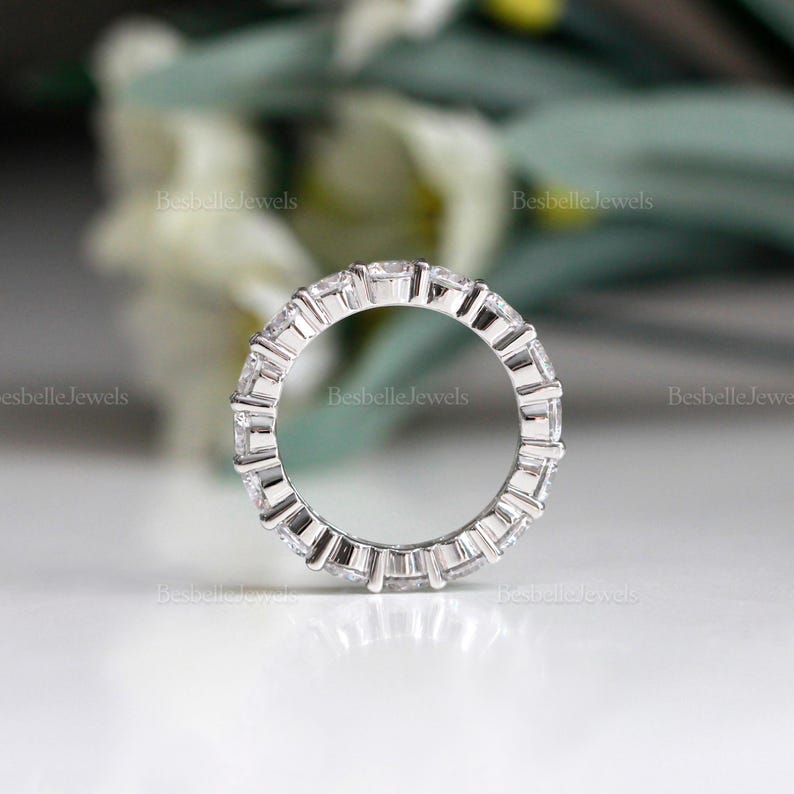 May include: A silver ring with a continuous row of square-cut, clear gemstones. The ring is set in a silver band and is displayed against a soft, blurred background. The ring is a piece of jewelry.