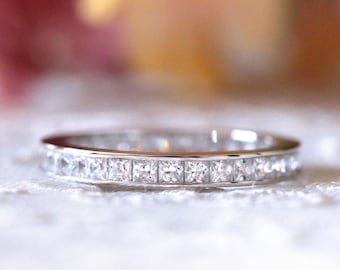 May include: A silver ring with a band of square-cut, clear gemstones. The ring is set on a white, textured surface, with a blurred background of pink and yellow hues. The ring is a piece of jewelry.