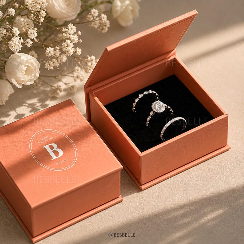以下が含まれることがあります： テラコッタ色の正方形のジュエリーボックス。蓋には白い文字で「BESBELLE」と書かれ、文字「B」が入った円形のロゴがあります。中には、黒いベルベットの台座の上に、透明な石があしらわれた3つの銀色の指輪が収められています。