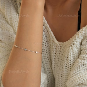 Puede incluir: Una delicada pulsera de plata con cuatro gemas redondas y transparentes, espaciadas uniformemente en una fina cadena. La pulsera se muestra en un brazo, con un suave suéter blanco en el fondo. La marca "Besbelle Jewels" es visible.