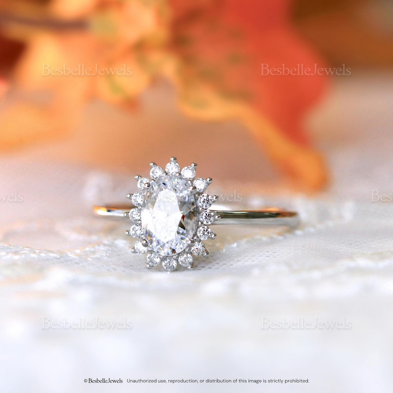 May include: A silver ring with a large, oval-cut, clear gemstone surrounded by smaller, round gemstones. The ring is set on a white lace surface, with the words "Besbelle Jewels" visible in the background.