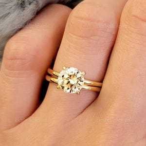 May include: A gold engagement ring with a large, round, clear diamond center stone. The ring is worn on a finger with a matching gold band.
