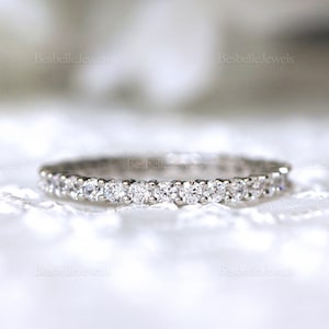 Op de afbeelding: Een zilverkleurige ring, bezet met talrijke kleine, heldere, fonkelende edelstenen. De ring wordt gepresenteerd op een wit, getextureerd oppervlak. De merknaam "Besbelle Jewels" is zichtbaar op de achtergrond.