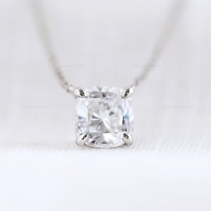 Puede incluir: Un collar con colgante de piedra preciosa transparente de corte cuadrado. La piedra está engastada en un metal plateado con cuatro garras. La cadena del collar también es plateada. El fondo es una tela blanca suave.