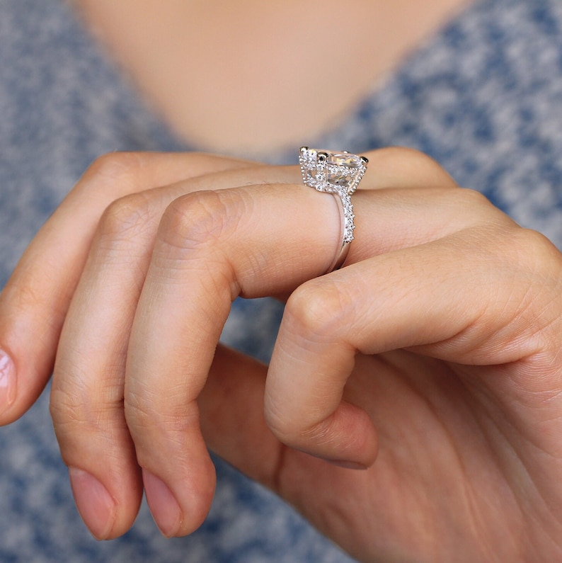May include: A silver engagement ring with a large, rectangular-cut center stone and a halo of smaller stones. The band is also adorned with small stones. The ring is on a person's finger, with a blue background.
