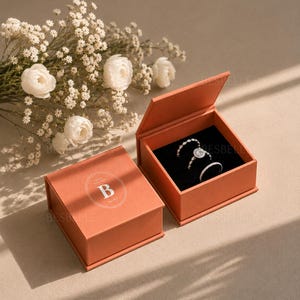 May include: Two square terracotta-colored jewelry boxes with hinged lids. One box is closed, displaying a white circular logo with the letter B. The other box is open, revealing a black velvet interior holding three silver-toned rings with stones.